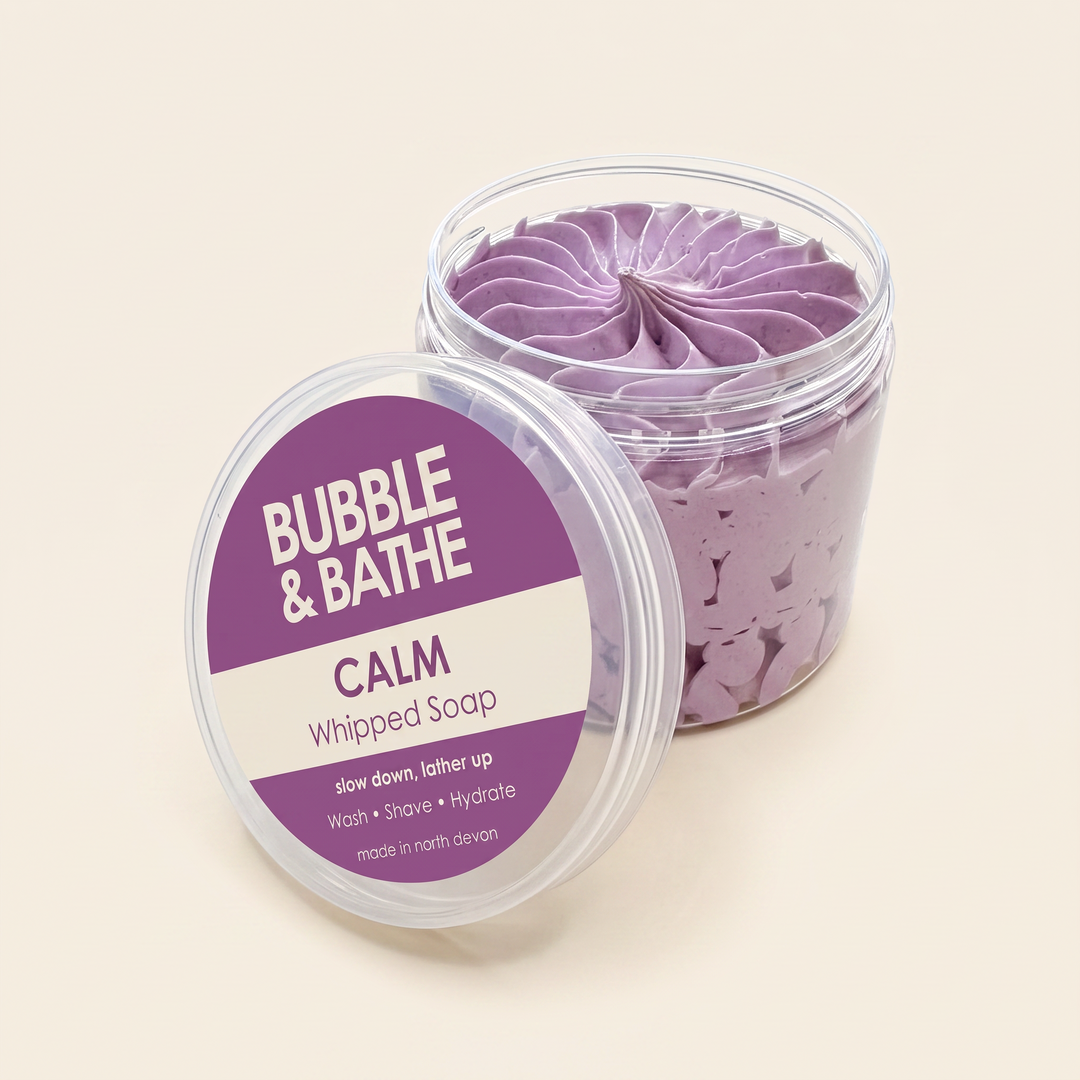 A clear plastic tub of whipped soap with its lid leaning beside it on a light beige background. The lid label reads 'BUBBLE & BATHE CALM Whipped Soap' with additional text below.