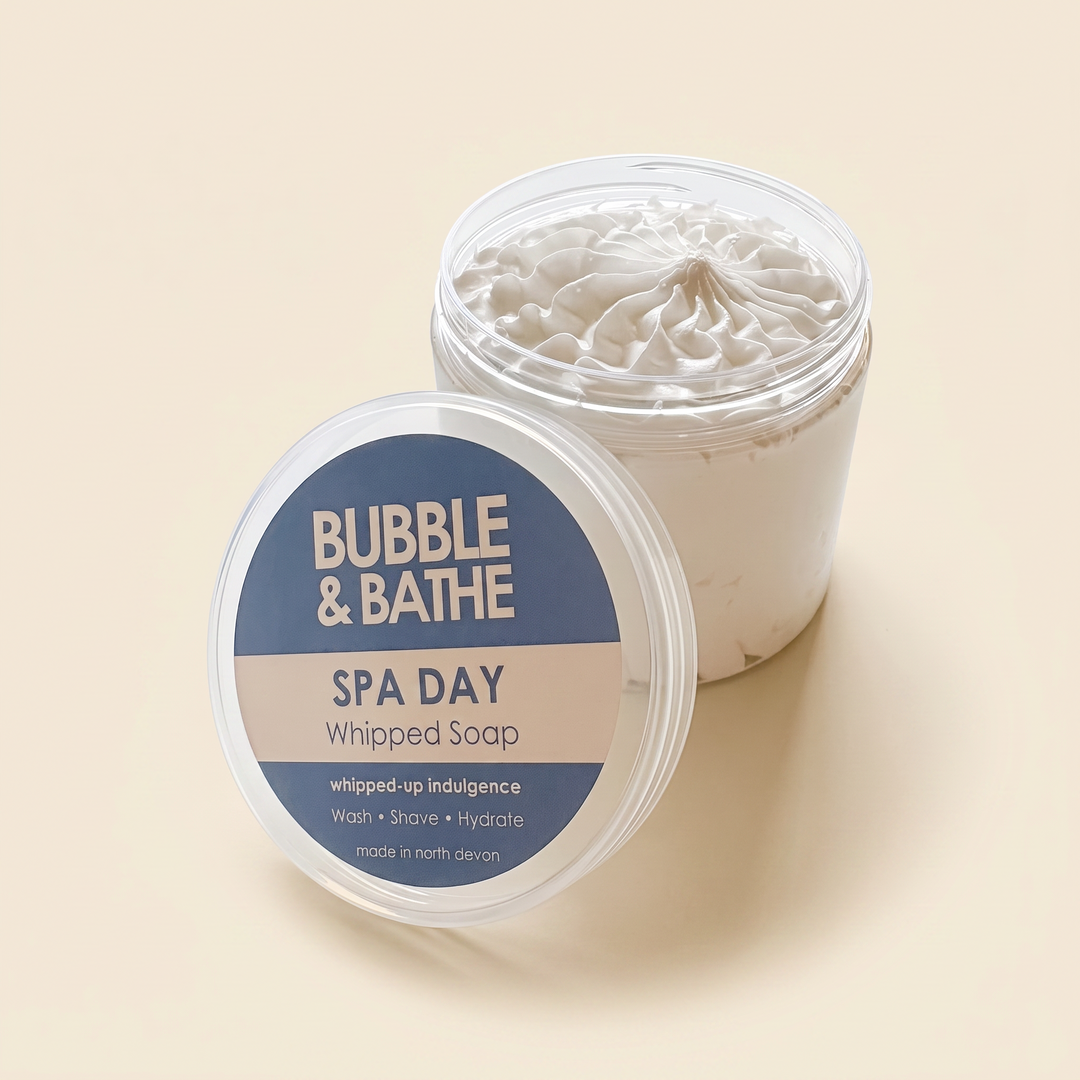 A clear plastic tub of whipped bath soap with its lid placed in front, showing the product and label on a warm beige background. The label reads 'BUBBLE & BATHE SPA DAY Whipped Soap'.